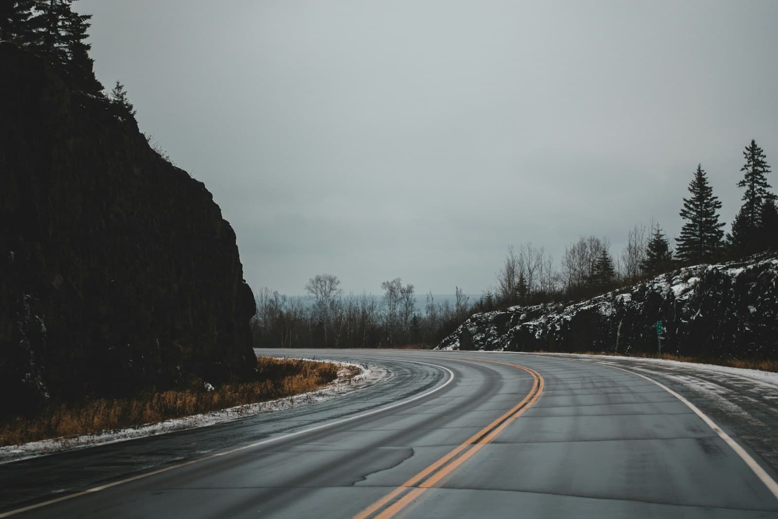 Wet curving two-lane highway through snow-patched hillsides and evergreens