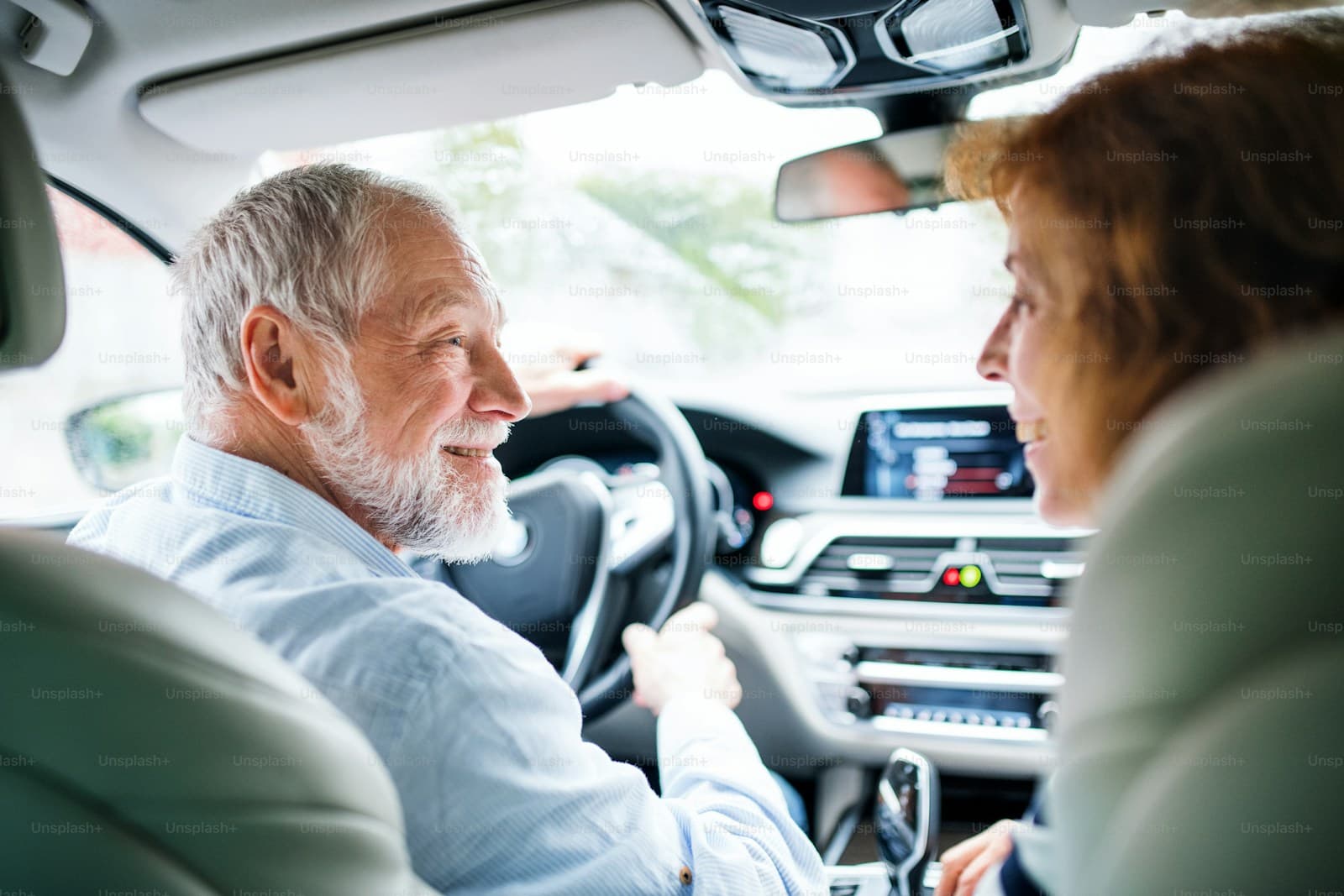 Smiling elderly man with gray hair and beard driving while talking with a front-seat passenger